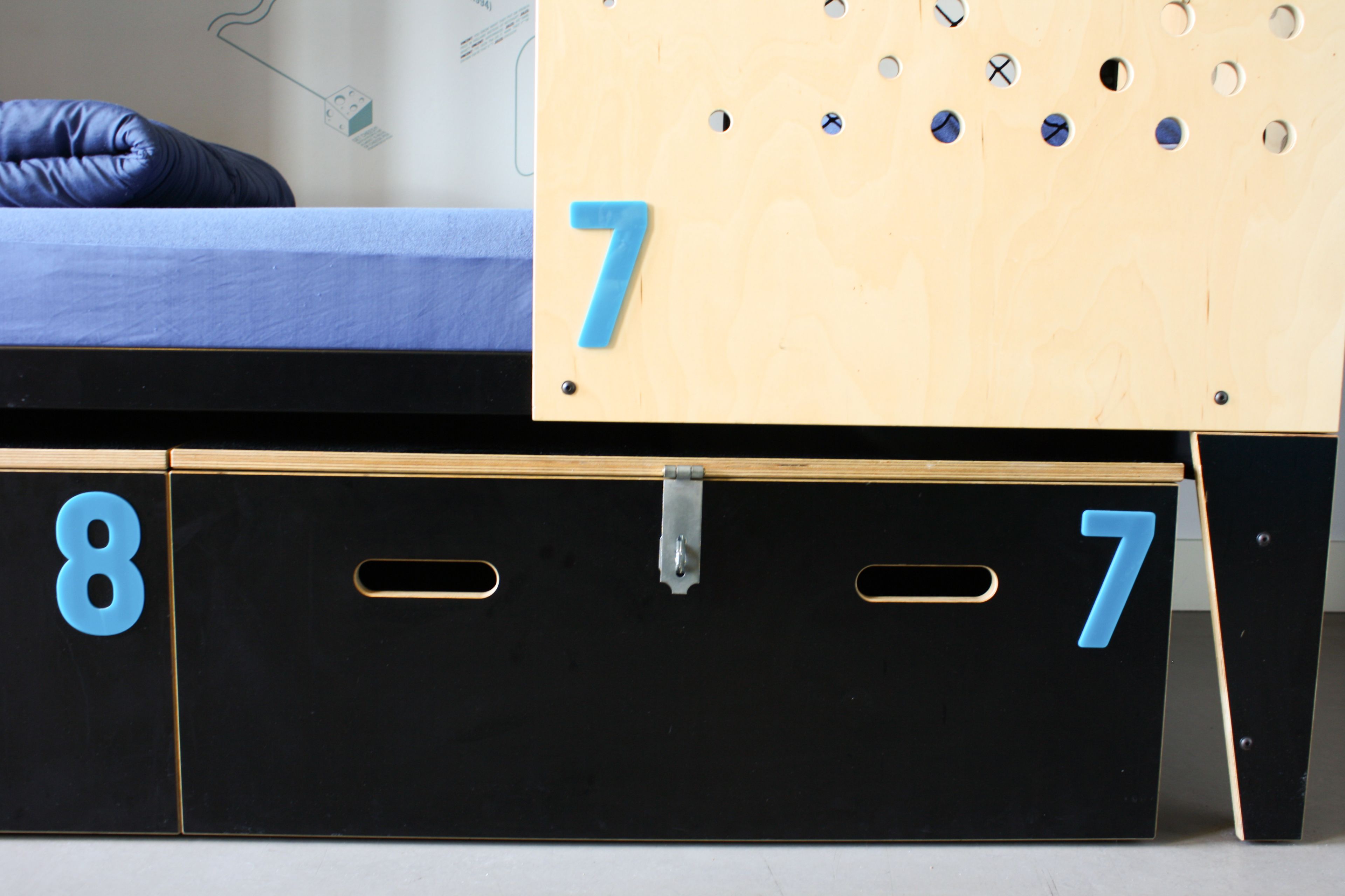 Ein Bett mit blauer Matratze, einem Holzkopfteil mit Löchern und einem schwarzen Stauraum mit blauen Zahlen.