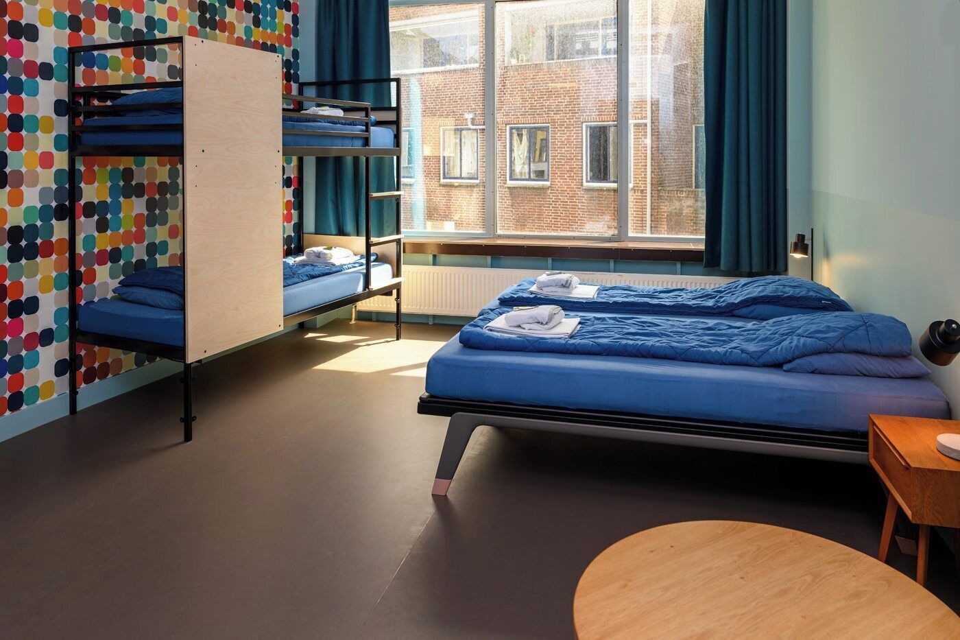 A room with bunk beds, a round table, and a view of a brick building through a window.