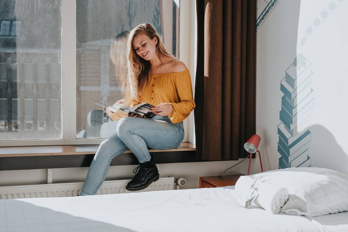 A person sitting on a window sill in a room with a bed, a lamp, and a wall with architectural drawings.