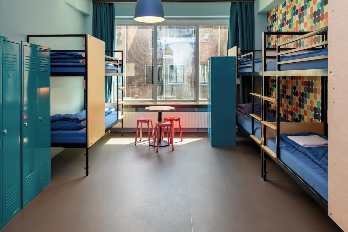 A dormitory room with bunk beds, a small table, and chairs, a window with curtains, and a radiator.