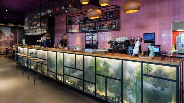 A bar area with a long counter, stools, shelves with bottles, and a refrigerator. - Stayokay Utrecht Centrum - Hostel (Utrecht)
