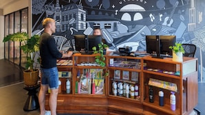 A man standing at a wooden desk with two computers, surrounded by shelves with travel brochures and bottles. - Stayokay Utrecht Centrum - Hostel (Utrecht)