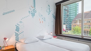 A hotel room with two beds, a nightstand with a lamp, a window with a view of buildings, and a wall with a cityscape mural. - Stayokay Utrecht Centrum - Hostel (Utrecht)