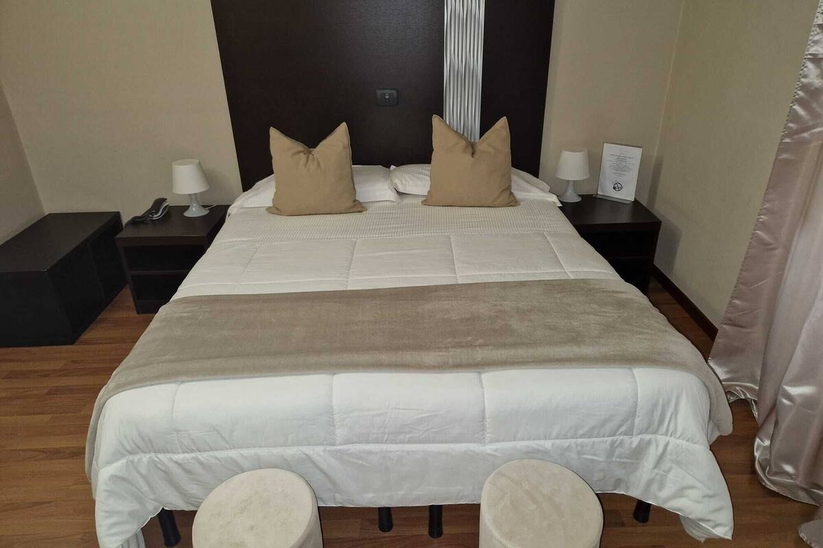 A neatly made bed with a white duvet and two beige pillows, flanked by two bedside tables with lamps and a phone.