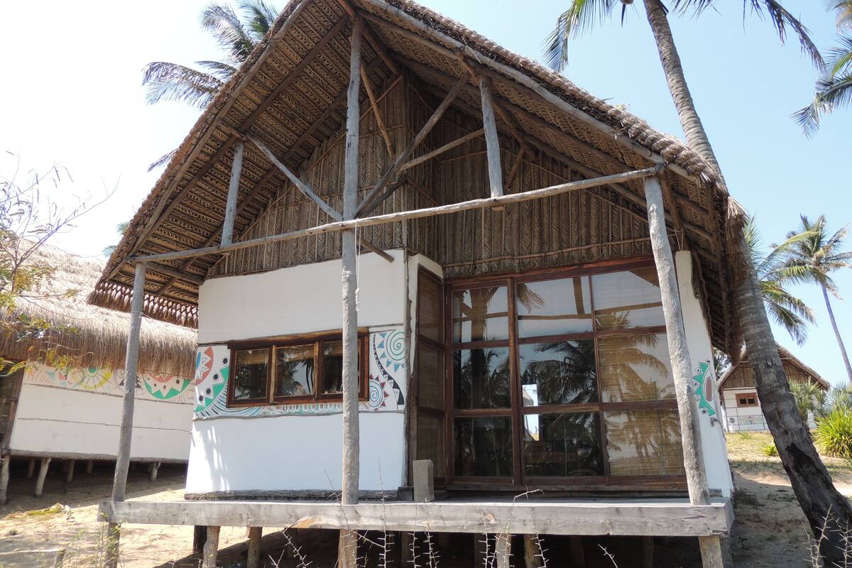 Una casa con techo de paja y ventanas amplias, rodeada de palmeras.