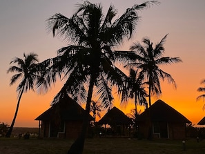 A sunset view with palm trees and thatched-roof huts.
