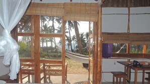 A room with a bed, a table, and a hammock. There are large windows with a view of palm trees and the beach. - Duna Lodge (Jangamo)