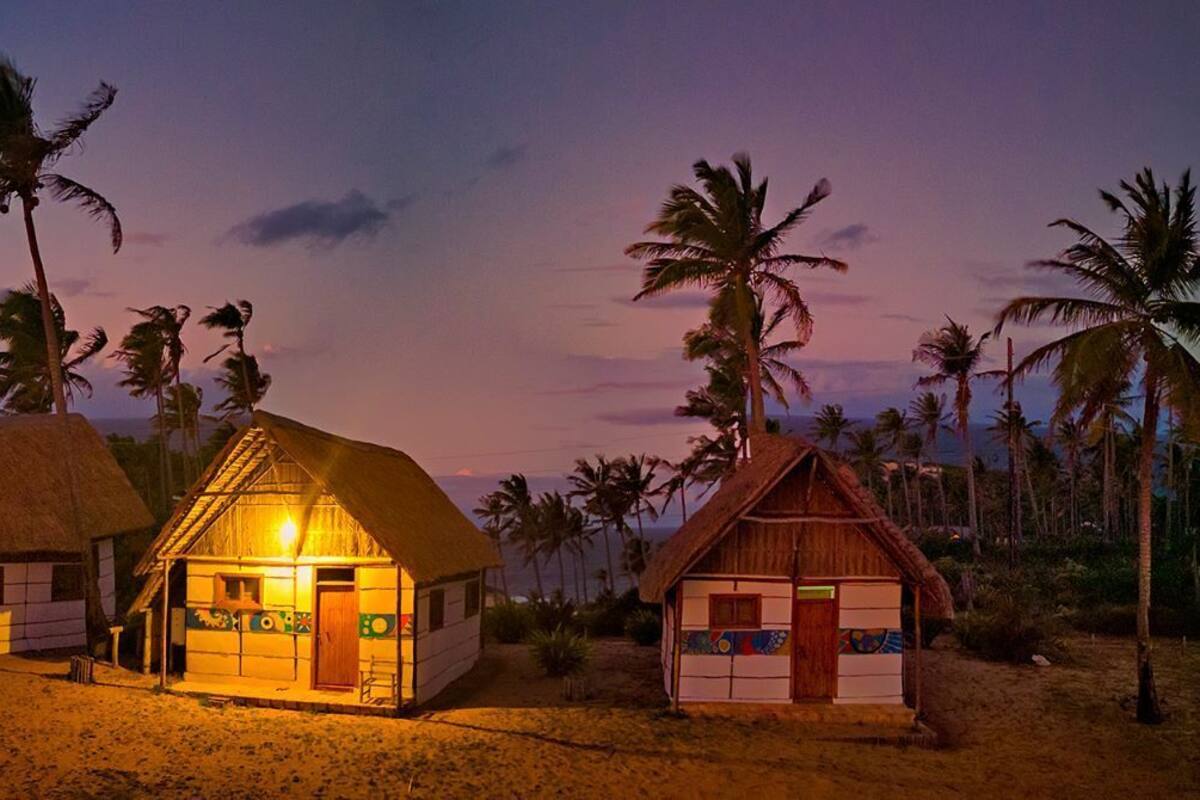 Una propiedad frente a la playa con cabañas de techo de paja, palmeras y un cielo al atardecer.