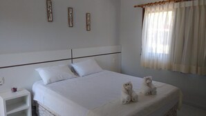 Een net opgemaakt bed met witte lakens, een nachtkastje met een rood voorwerp en een raam met dunne gordijnen.
