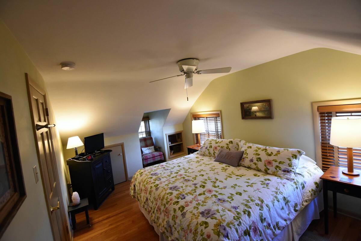 A bedroom with a bed, a ceiling fan, a desk with a lamp, a chair, a TV, and a window with blinds.