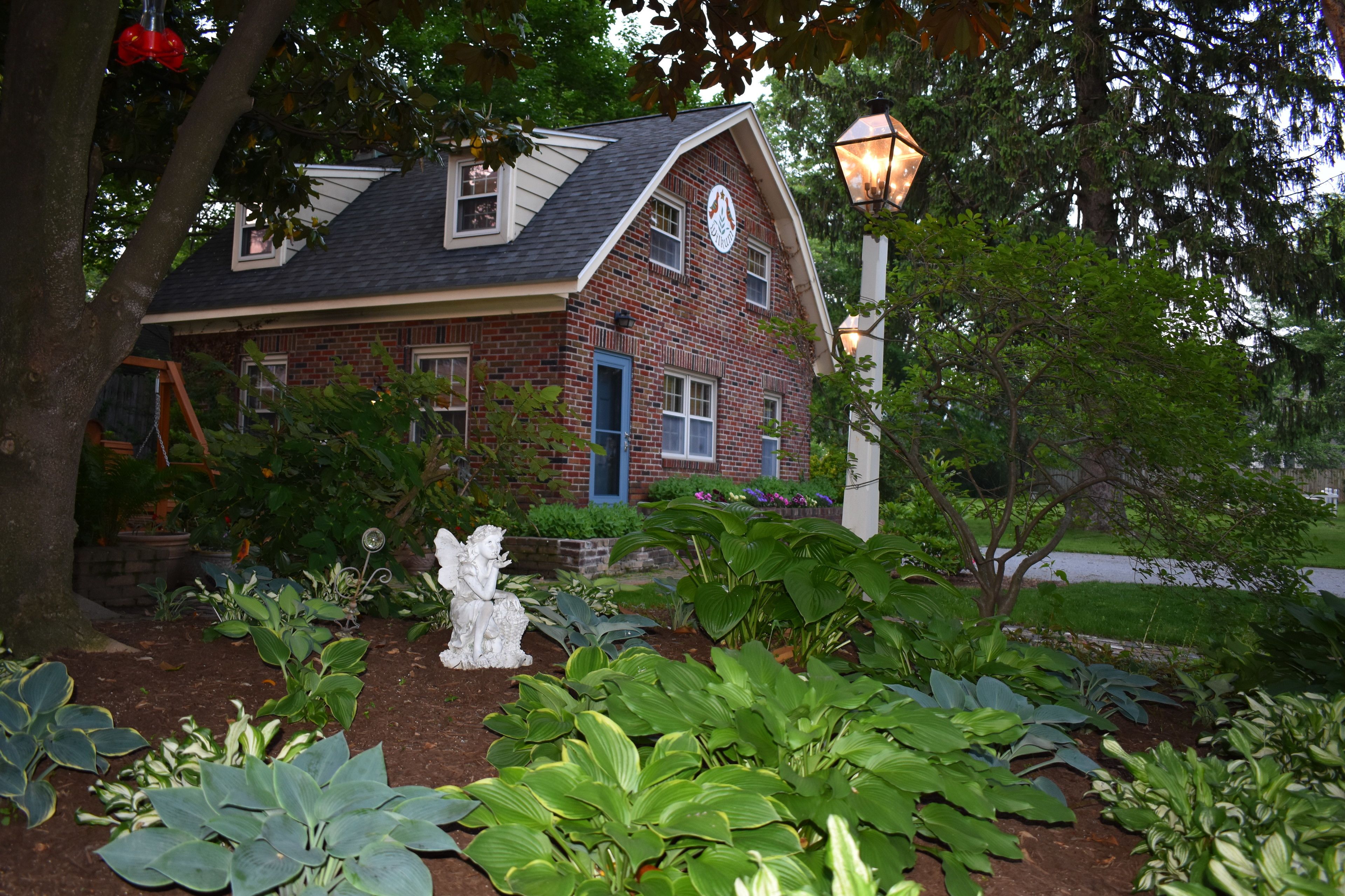 A brick house with a blue door and windows, surrounded by a garden with various plants and a white statue.