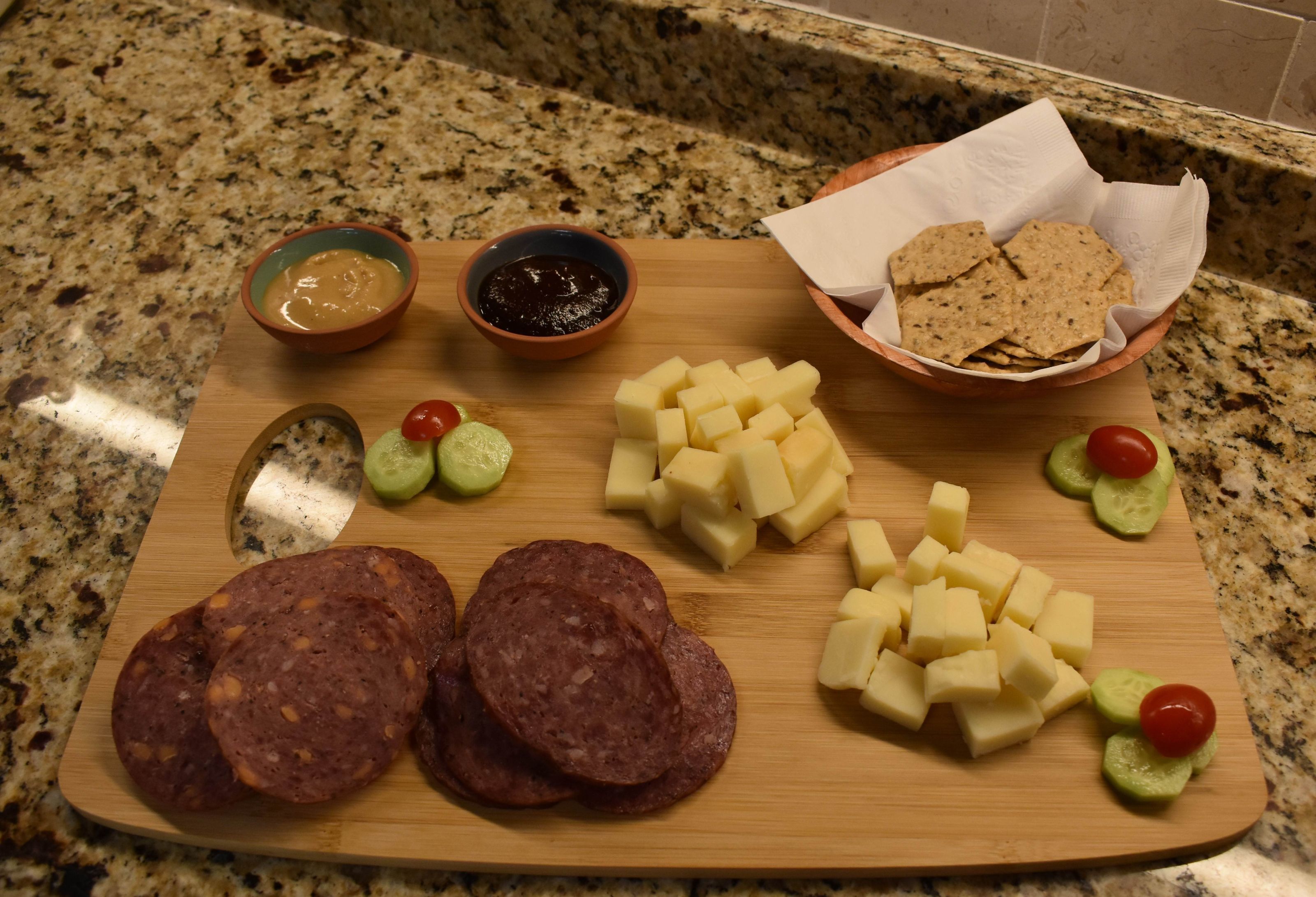 A wooden cutting board with sliced salami, cheese, cucumber slices, and cherry tomatoes on a granite countertop.