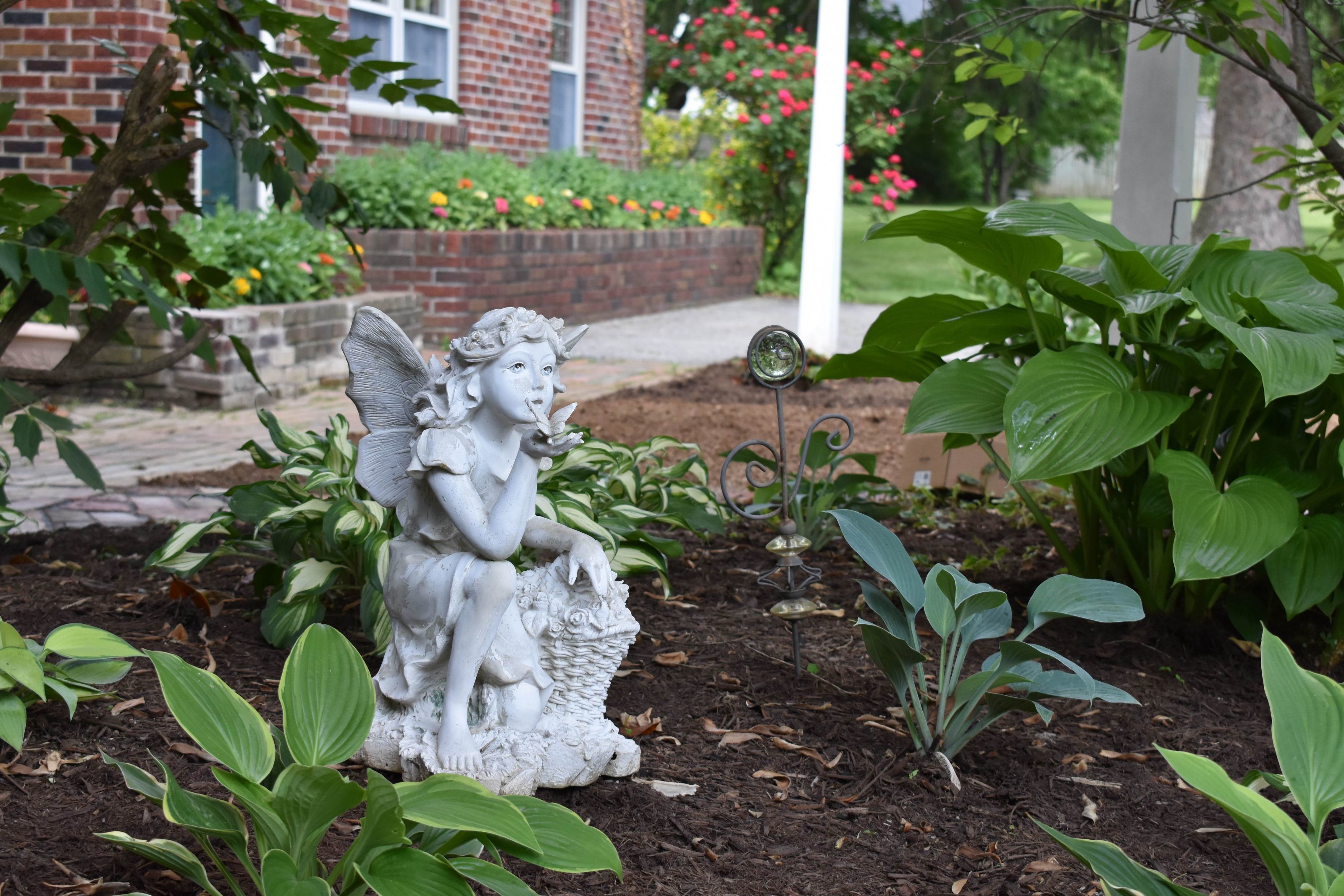 A garden with a statue of a figure holding a basket, surrounded by various plants and a brick building in the background.