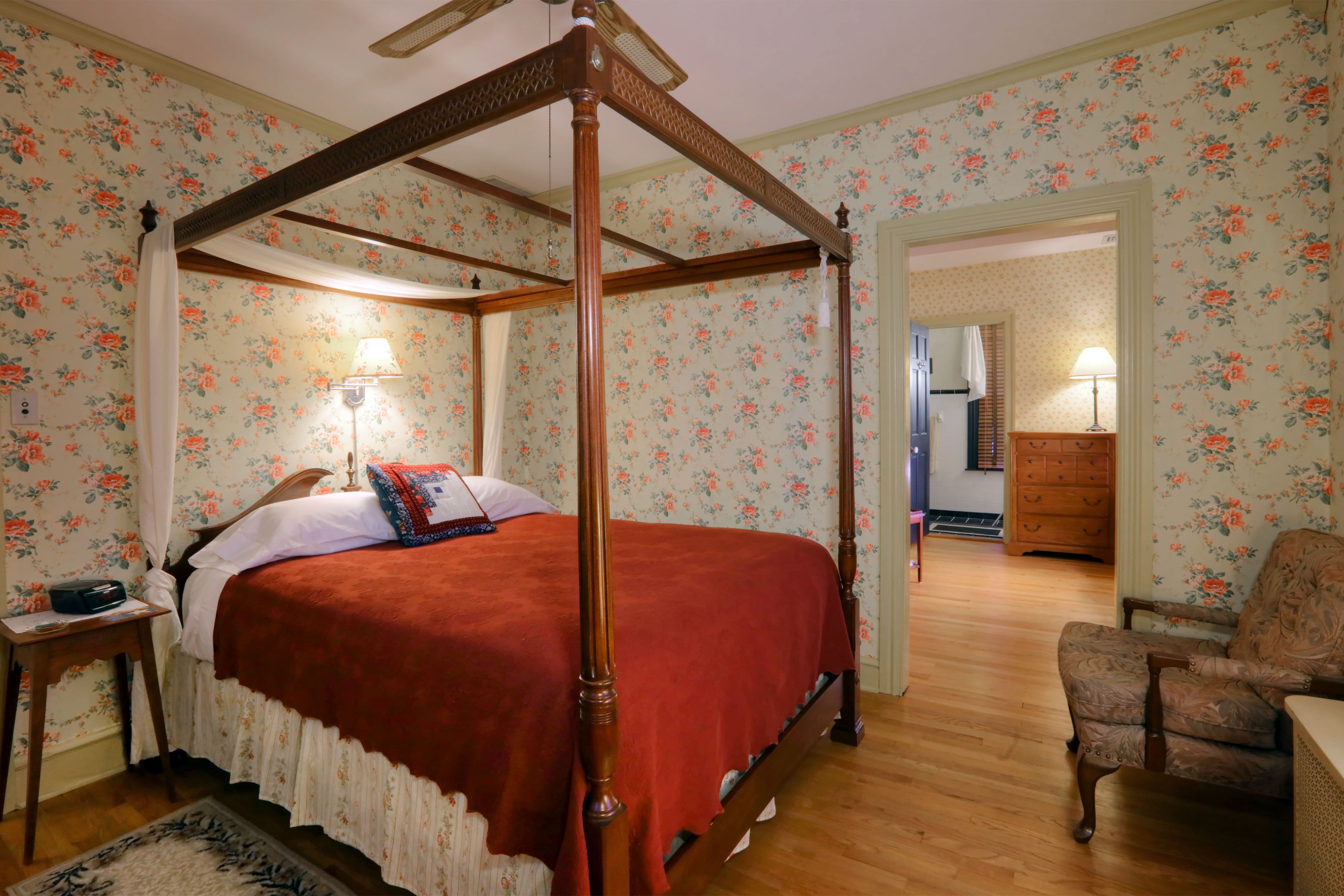 A four-poster bed with a red bedspread, a wooden nightstand with a lamp, and a floral-patterned chair.