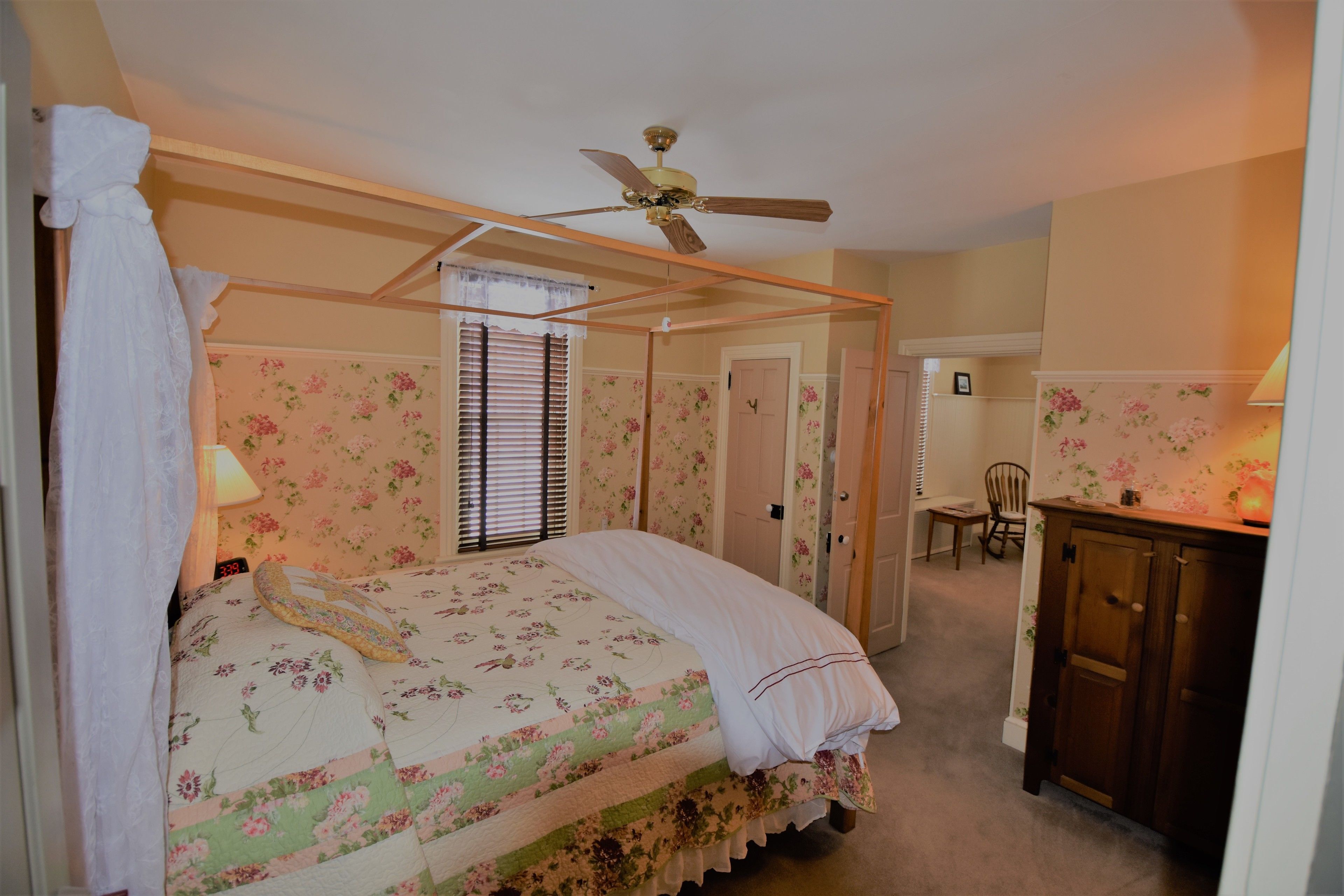 Quarto com cama de dossel, papel de parede floral, ventilador de teto e um armário de madeira.
