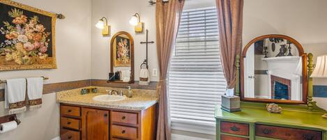 Un baño con tocador de madera, un espejo grande, un cuadro y una ventana con cortinas.