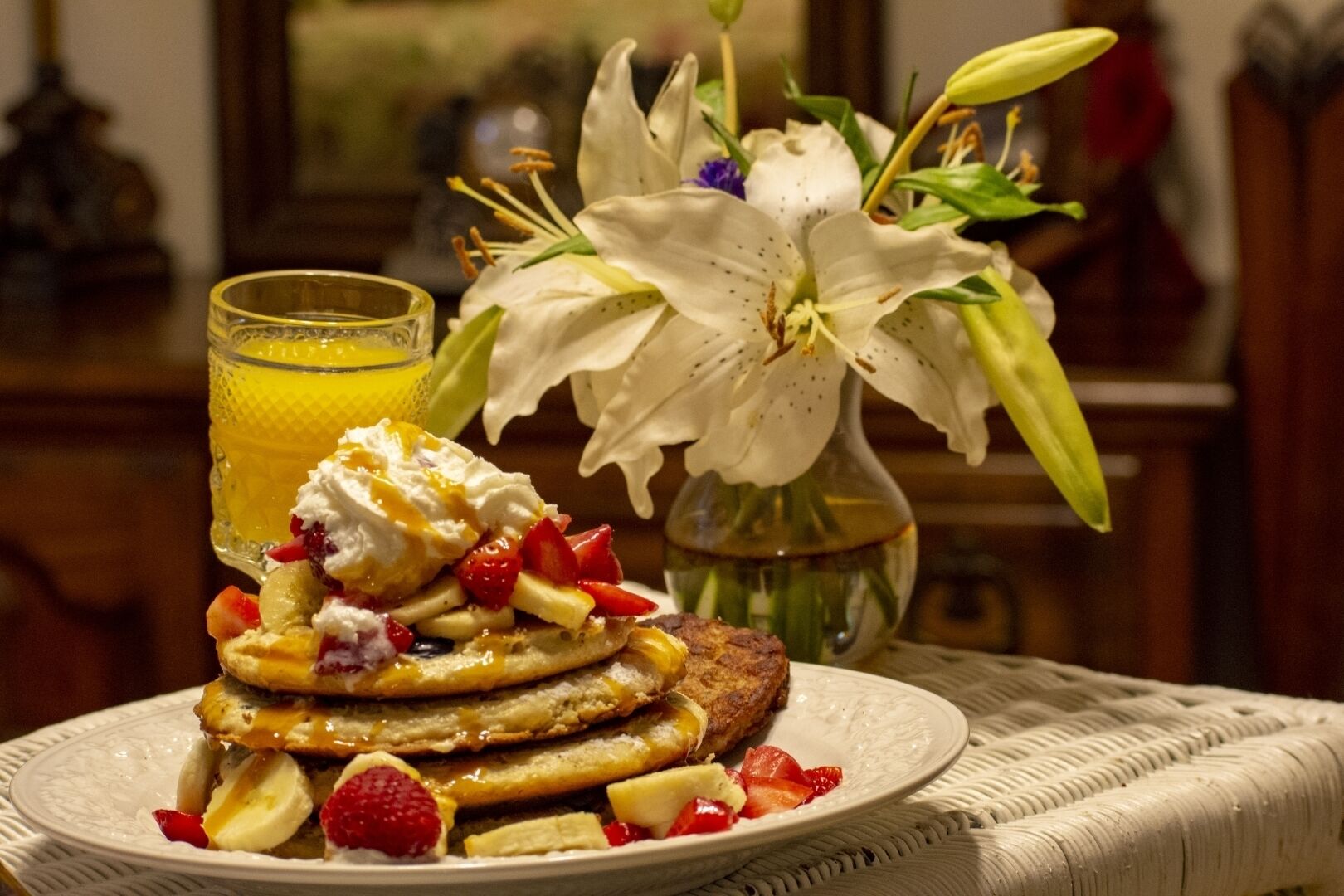 A plate of pancakes with fruit and whipped cream, a glass of orange juice, and a vase of white lilies.