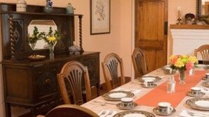 A dining room with a long table set for a meal, wooden chairs, a dark wooden cabinet, and a fireplace.