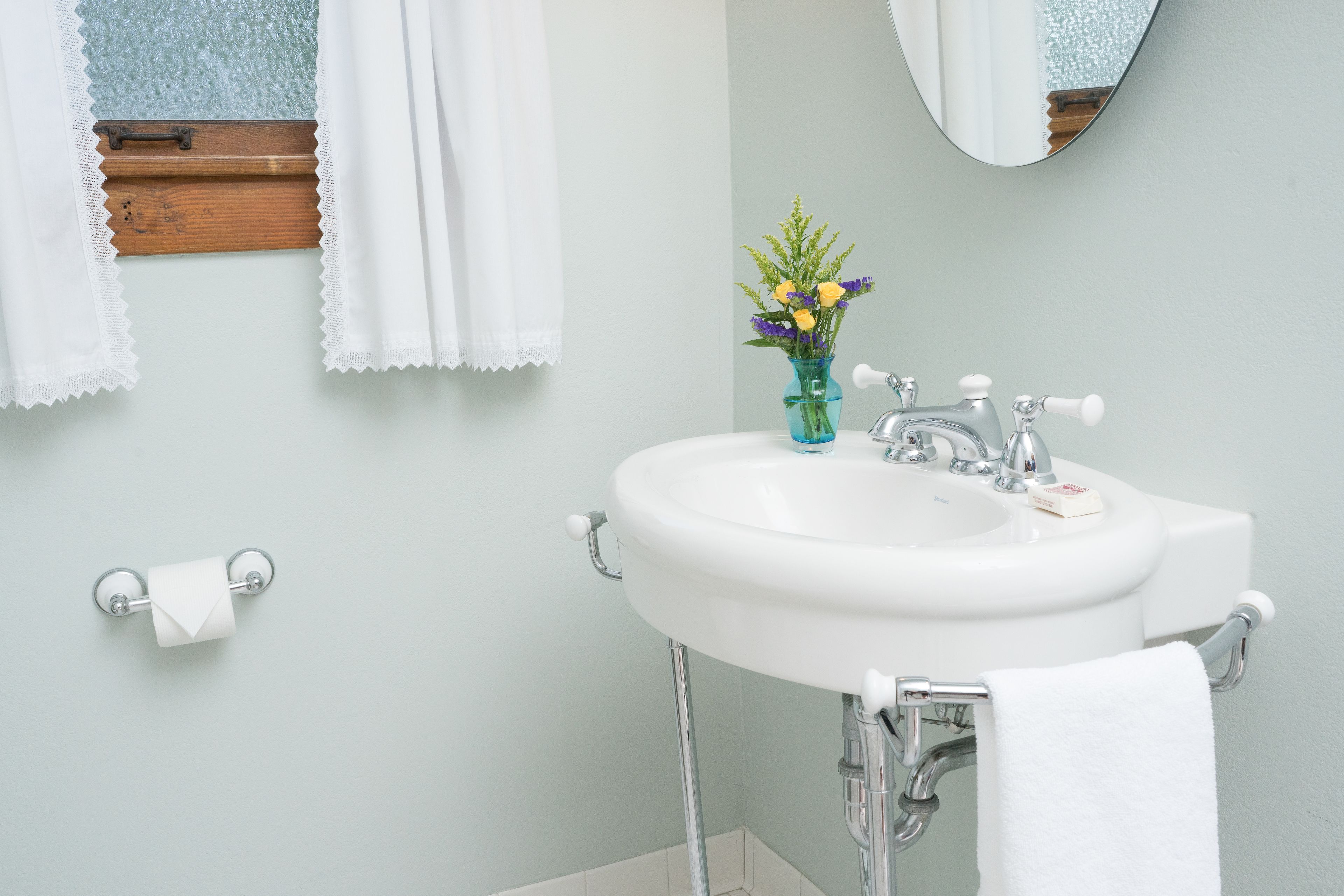 A bathroom with a white sink, a mirror, a window with lace curtains, and a vase with flowers.