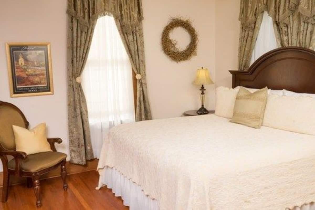 A bedroom with a large bed, a wooden chair, a nightstand with a lamp, and a framed picture on the wall.