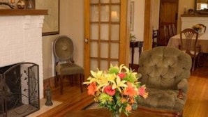 A living room with a white brick fireplace, a wooden floor, a grey armchair, and a vase of flowers.