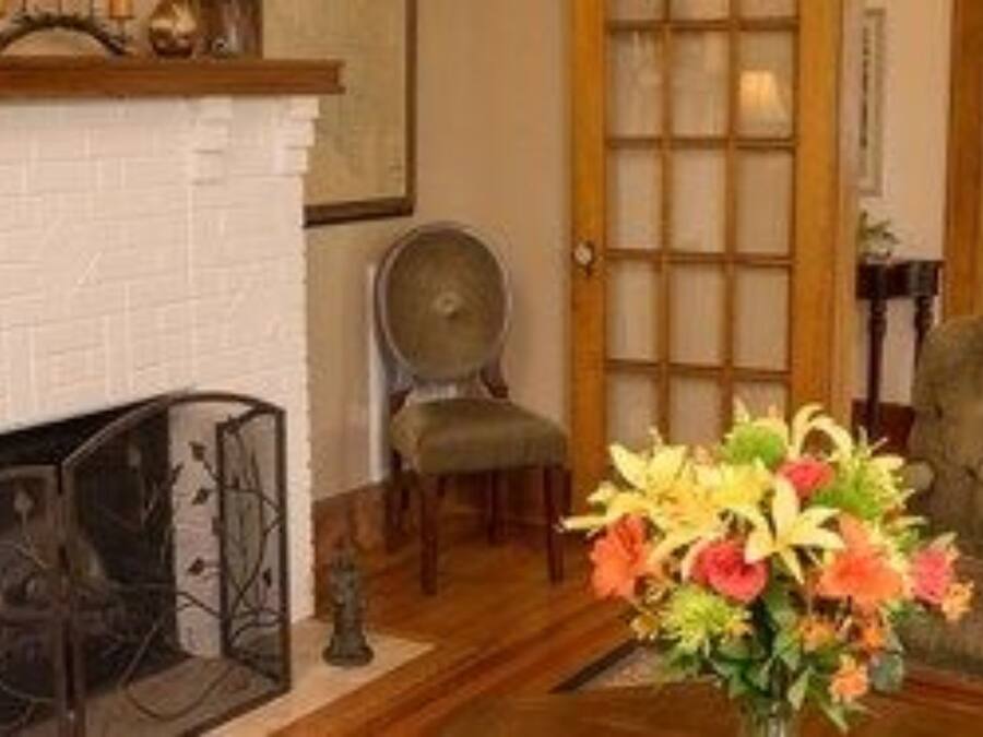 A living room with a white brick fireplace, a wooden floor, a grey armchair, and a vase of flowers.
