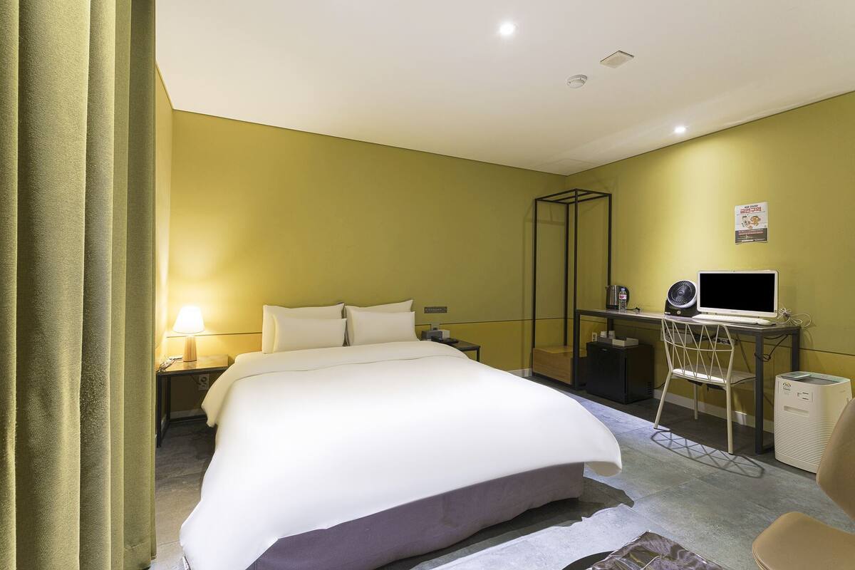 A hotel room with a bed, a desk with a computer, a chair, a mirror, a lamp, and a small table.