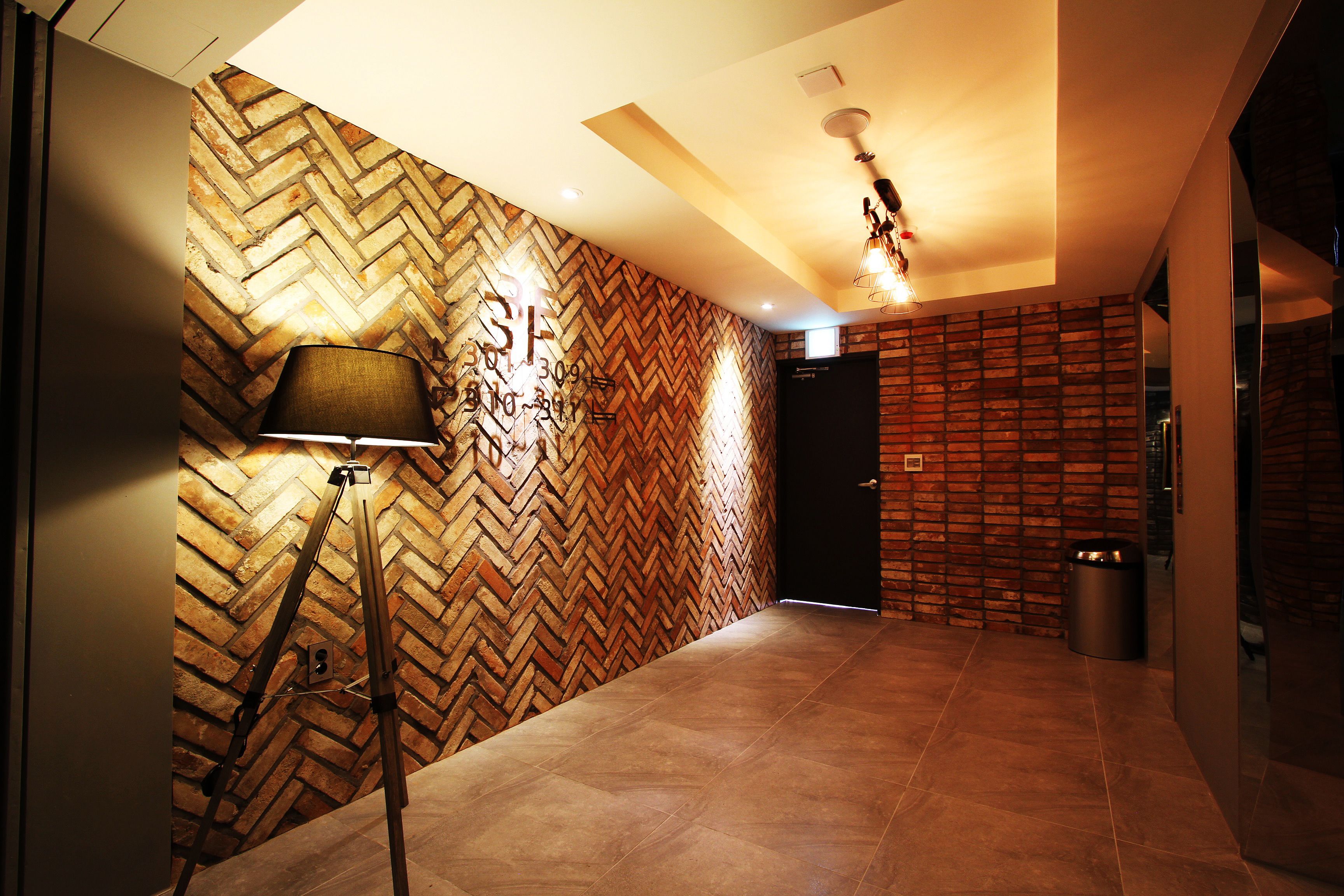 A hallway with a herringbone patterned wall, a lamp, and a trash bin.