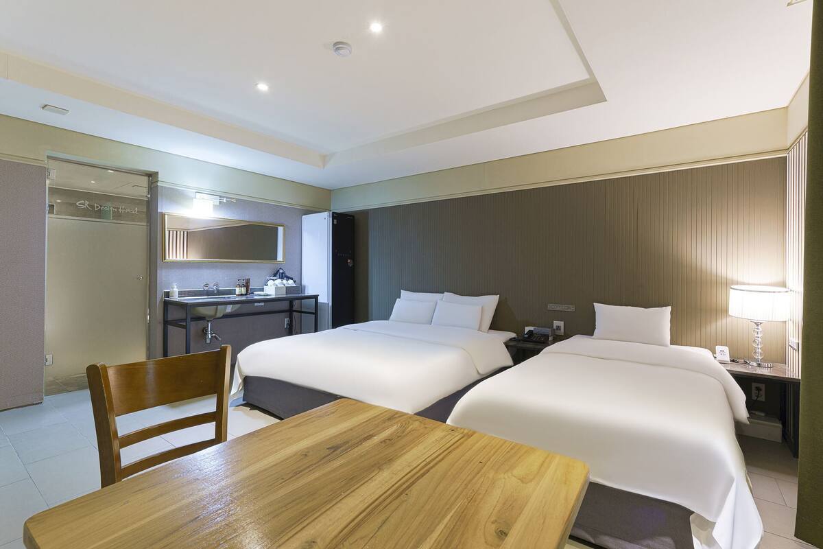 A hotel room with two beds, a wooden table, a desk with a mirror, a refrigerator, and a bathroom with a shower.
