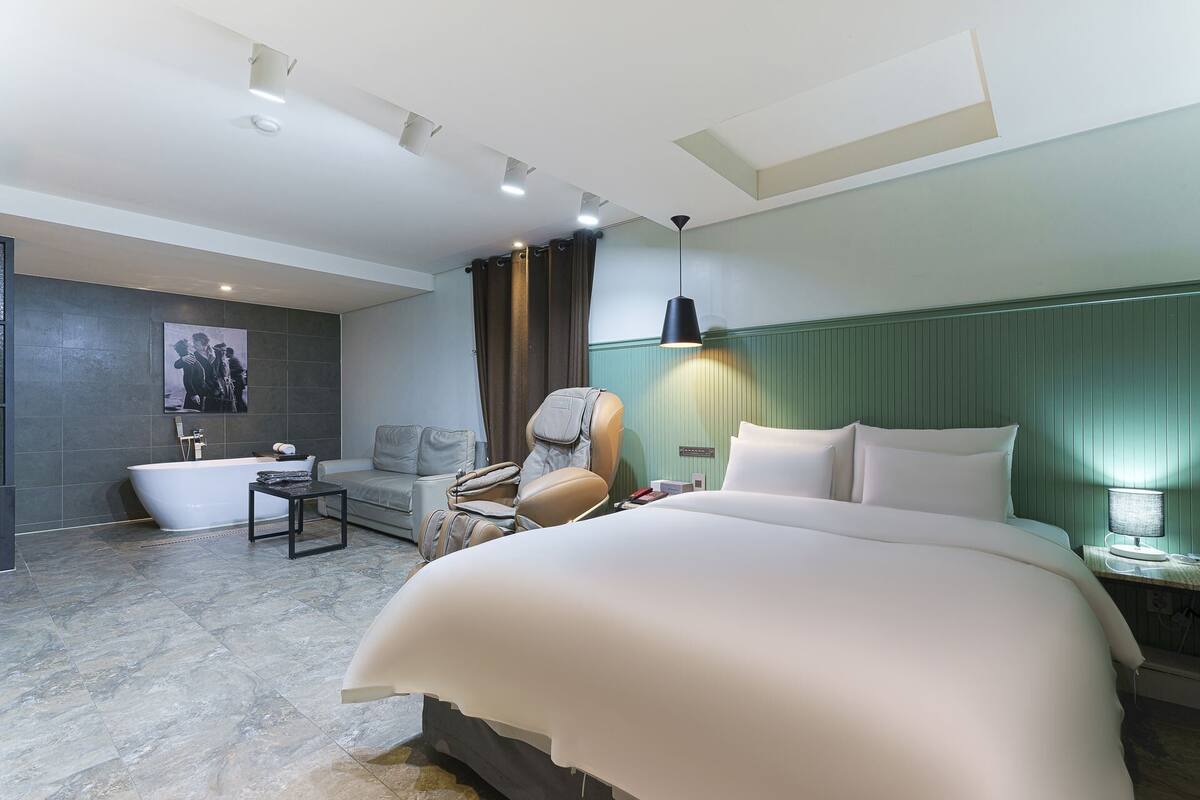 A hotel room with a large bed, a sitting area with a sofa and armchair, a bathtub, and a small table.