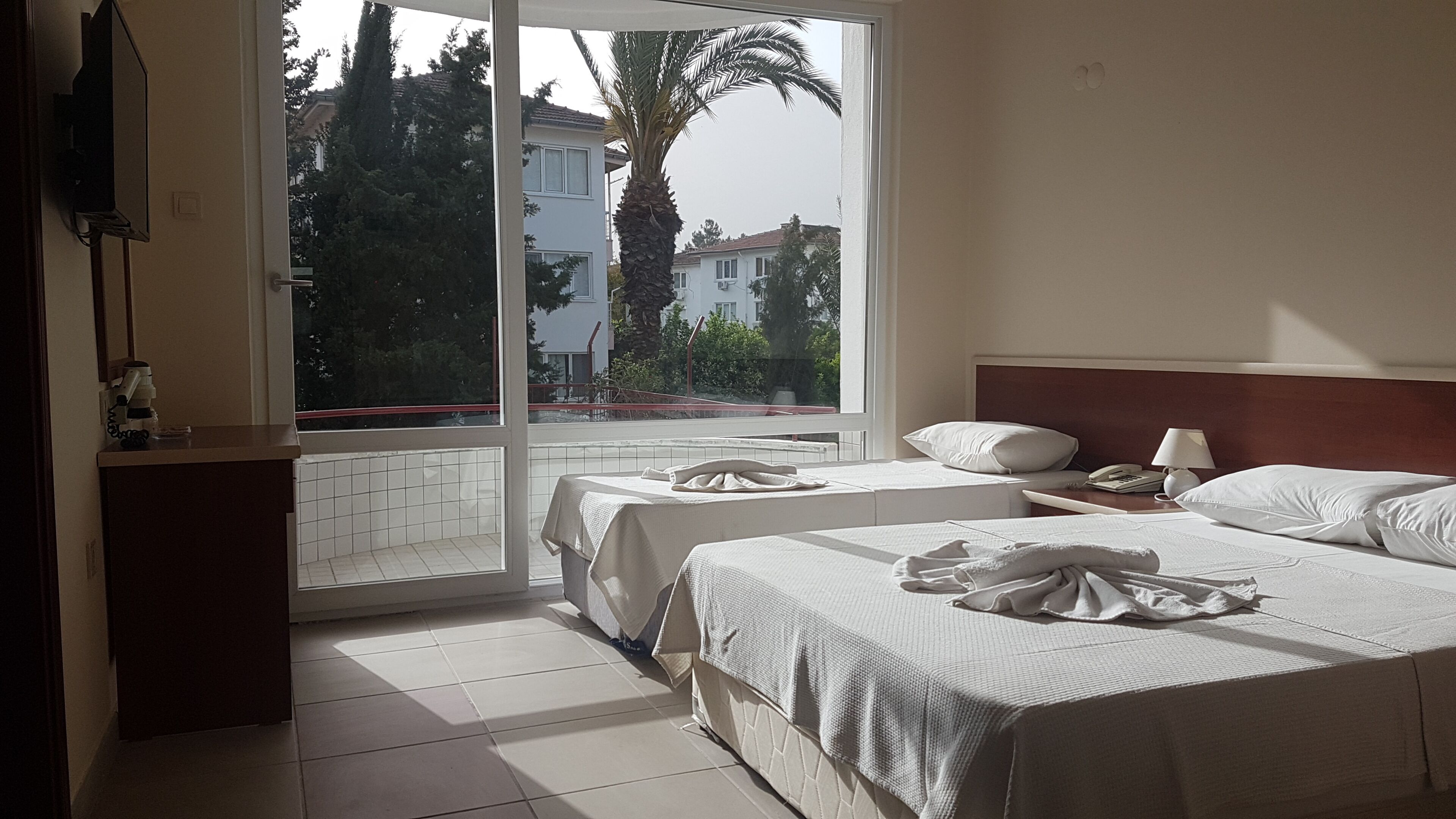 Habitación de hotel con dos camas, una ventana que ofrece vistas a edificios y árboles, una mesita de noche con lámpara y un escritorio pequeño con teléfono.