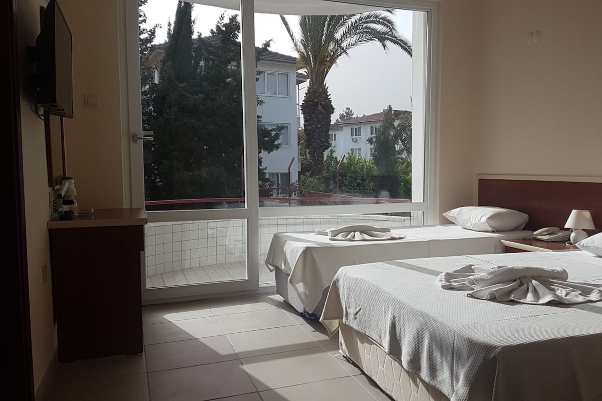 Habitación de hotel con dos camas, una ventana que ofrece vistas a edificios y árboles, una mesita de noche con lámpara y un escritorio pequeño con teléfono.