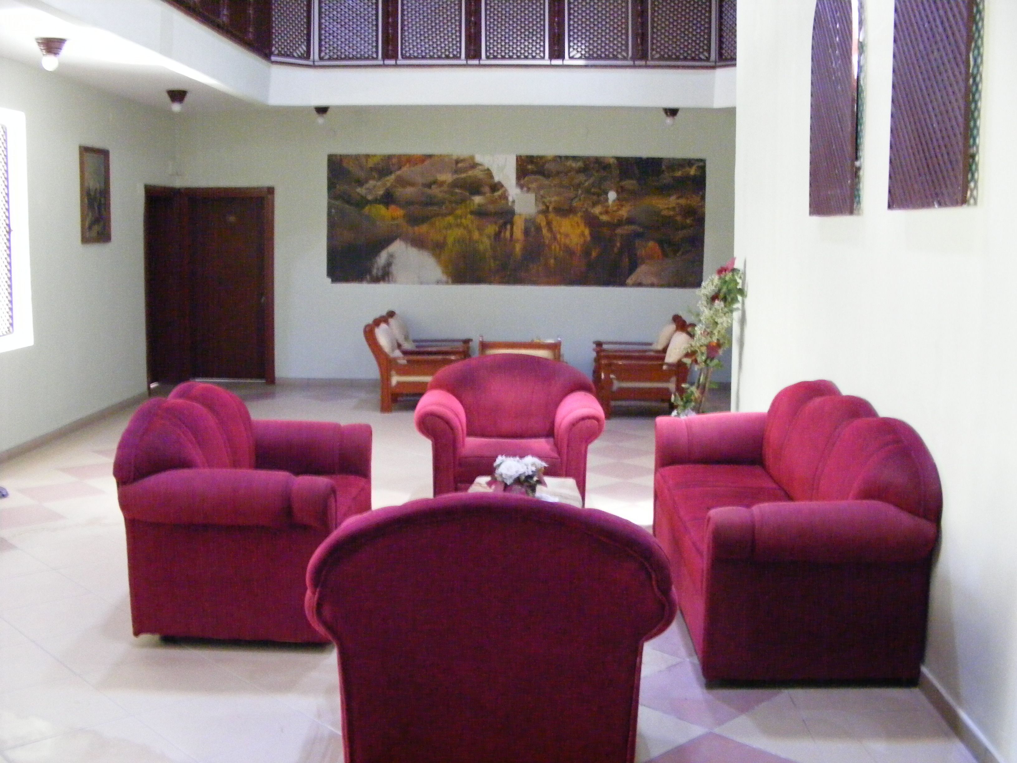 A spacious lobby with a high ceiling and a large painting on the wall. There are three plush red sofas arranged in a semi-circle, a wooden bench, and a small table with a vase of flowers.