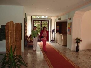 Un vestíbulo de hotel con alfombra roja, un mostrador de recepción de madera y un amplio ventanal con vistas a la vegetación.