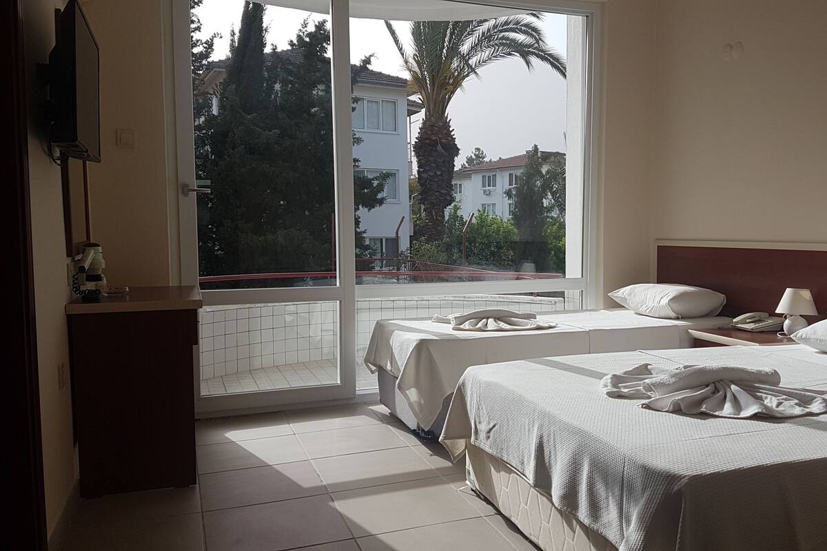 Habitación de hotel con dos camas, una ventana que ofrece vistas a edificios y árboles, una mesita de noche con lámpara y un escritorio pequeño con teléfono.