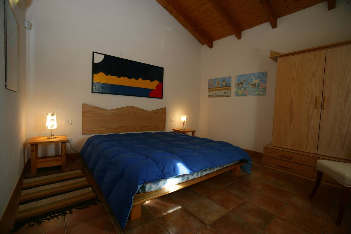 A bedroom with a wooden bed, two bedside lamps, a wooden headboard, a large painting above the bed, and a wooden wardrobe.
