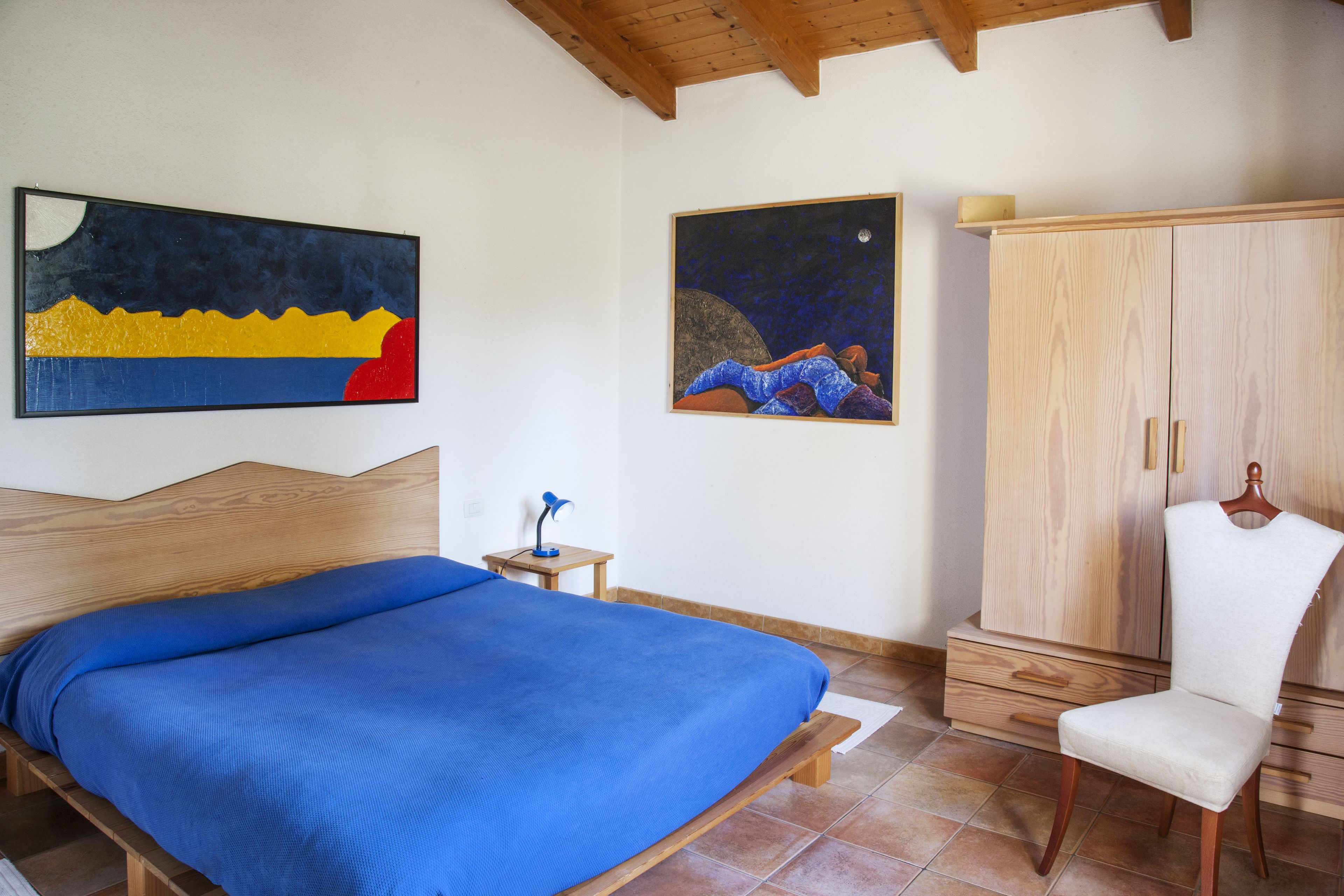 Un dormitorio con cama, mesita de noche, silla, armario y dos cuadros en la pared.