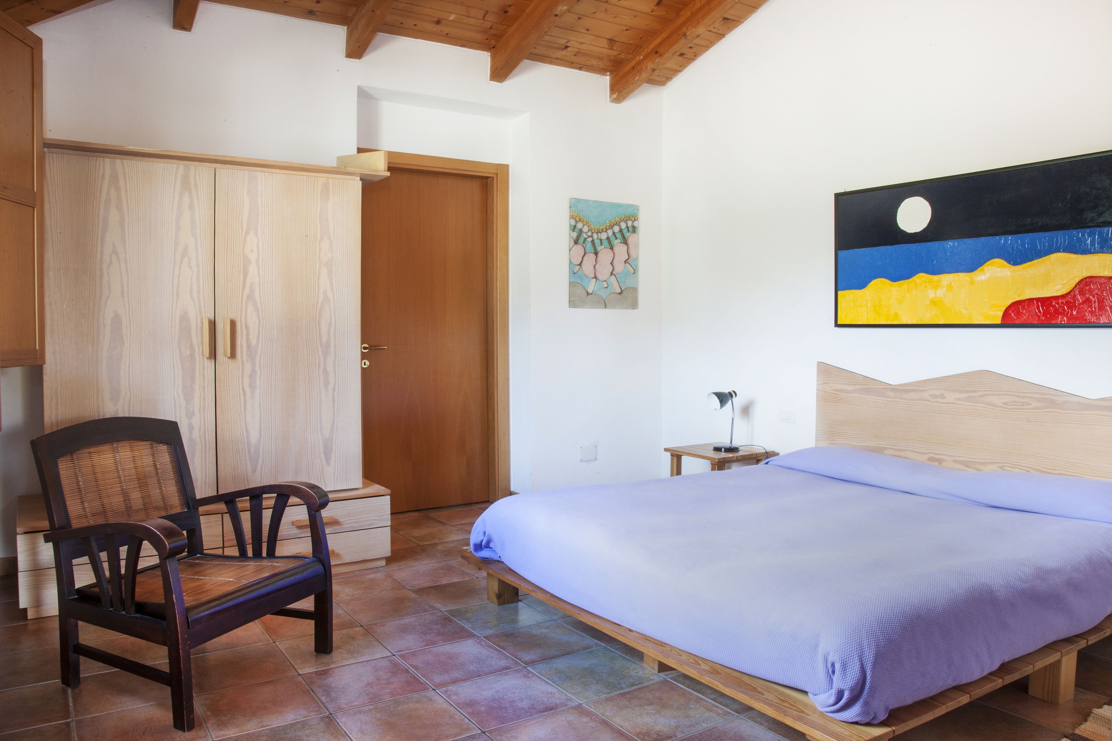 A bedroom with a bed, a wooden chair, a wardrobe, and a painting on the wall.