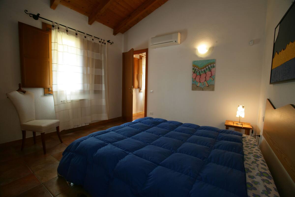 A bedroom with a large bed, a chair, a door, an air conditioning unit, a wall-mounted lamp, and a painting.