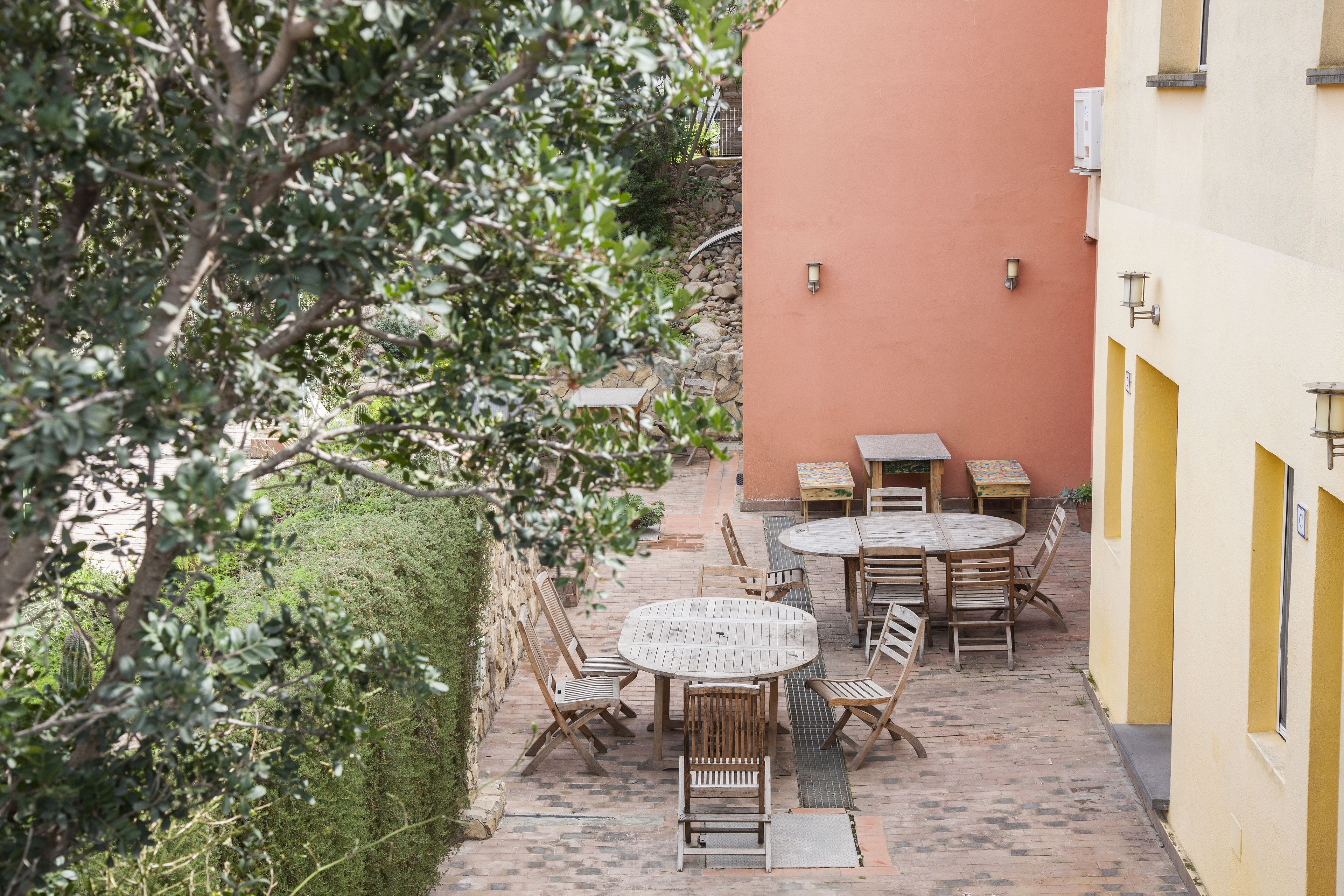Un espace extérieur aménagé en terrasse, avec des tables et des chaises rondes en bois, entouré d’un petit muret en pierre et de verdure.
