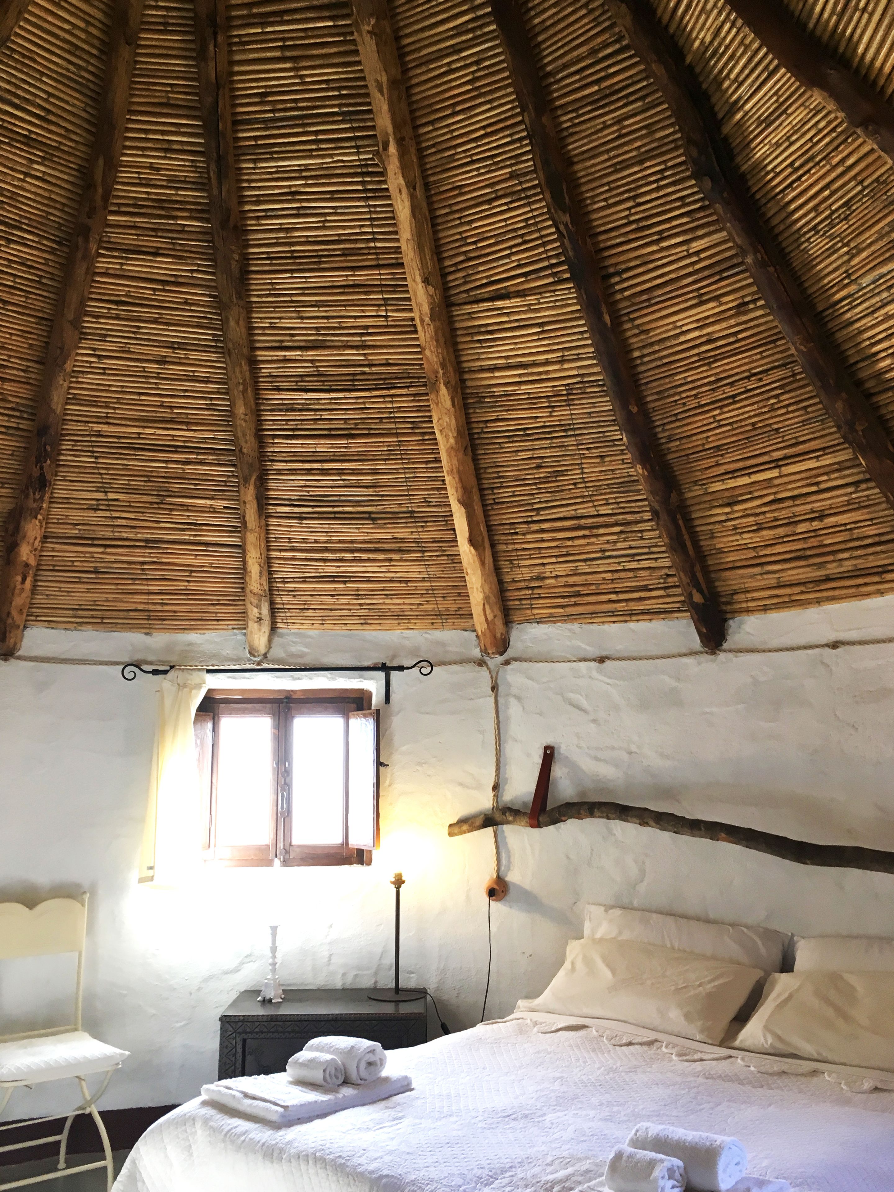 Una stanza rustica con tetto di paglia, un letto con cuscini, un comodino con una lampada e una finestra che fa entrare luce naturale.