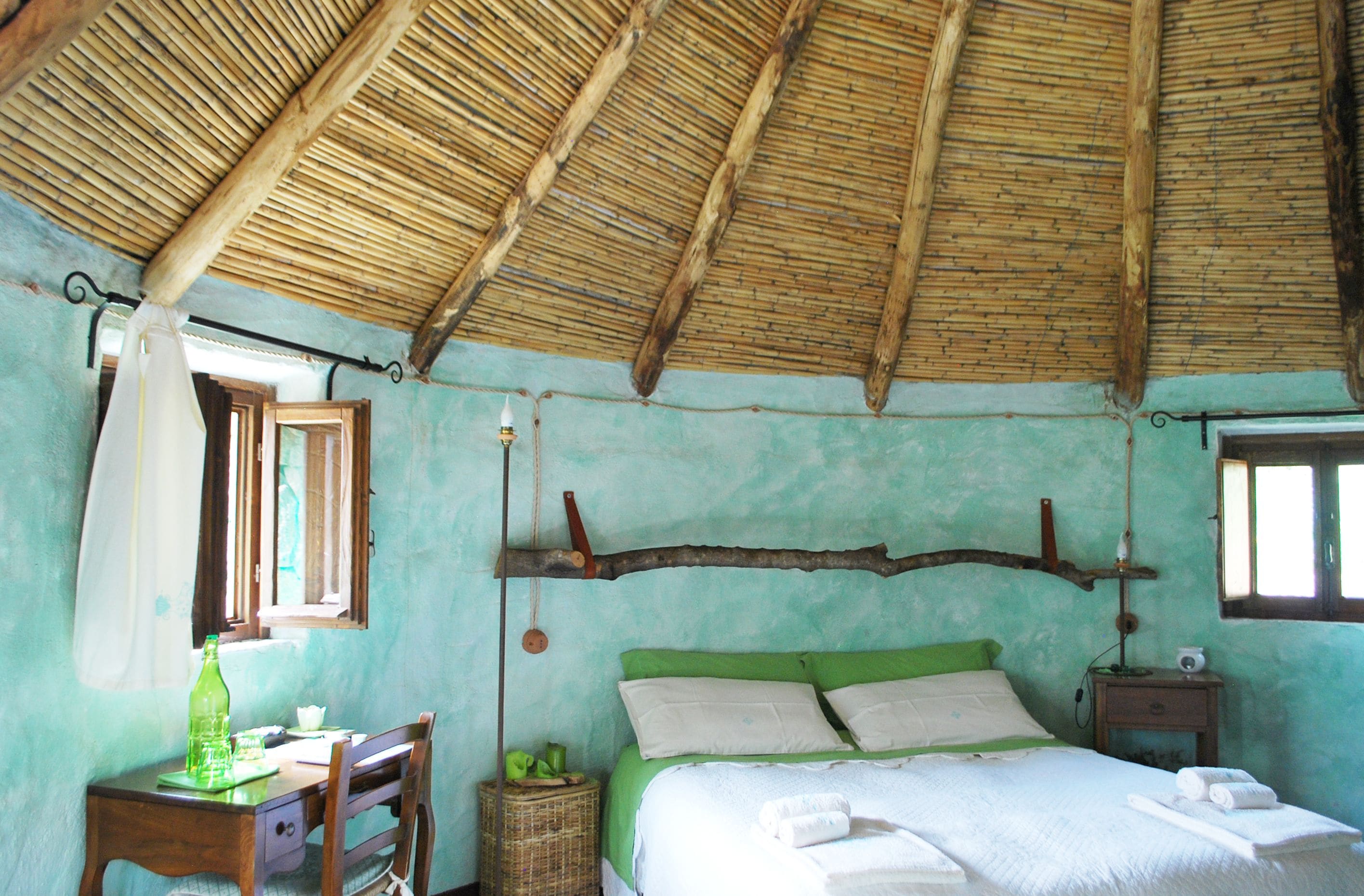Une pièce rustique avec un toit de chaume, un lit avec une literie verte, un bureau en bois et une chaise en osier.