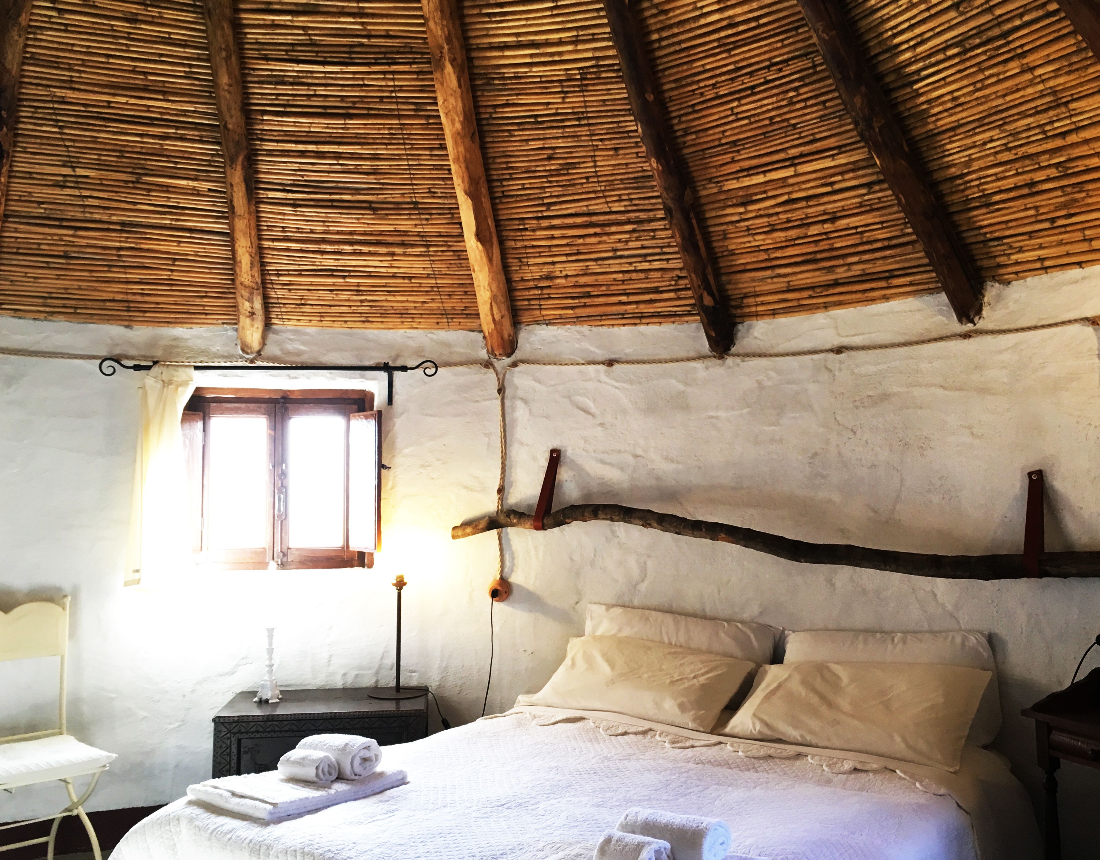 Une pièce rustique avec un toit de chaume, un lit recouvert de draps blancs, une petite table avec une lampe et une fenêtre avec des rideaux.