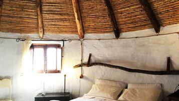 Une pièce rustique avec un toit de chaume, un lit recouvert de draps blancs, une petite table avec une lampe et une fenêtre avec des rideaux.