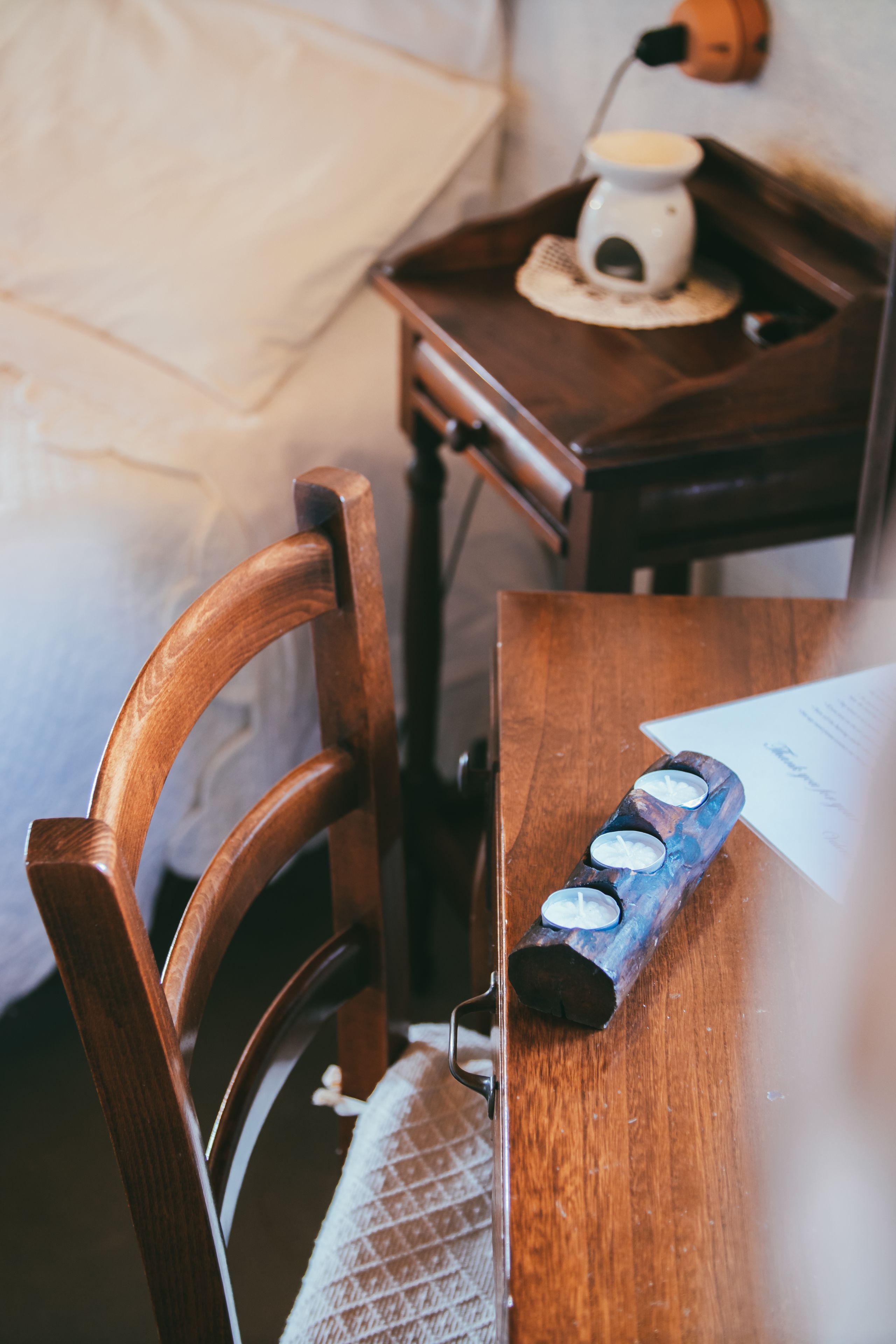 A wooden chair with a woven seat cushion, a wooden table with a tray holding a white object and a blue object, and a small white table lamp on a wooden nightstand.
