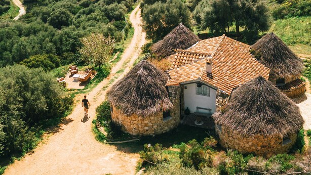 A rustic house with a tiled roof surrounded by thatched-roof structures, set in a lush, green landscape with a dirt path leading to the house. - Essenza Sardegna (Torpe)