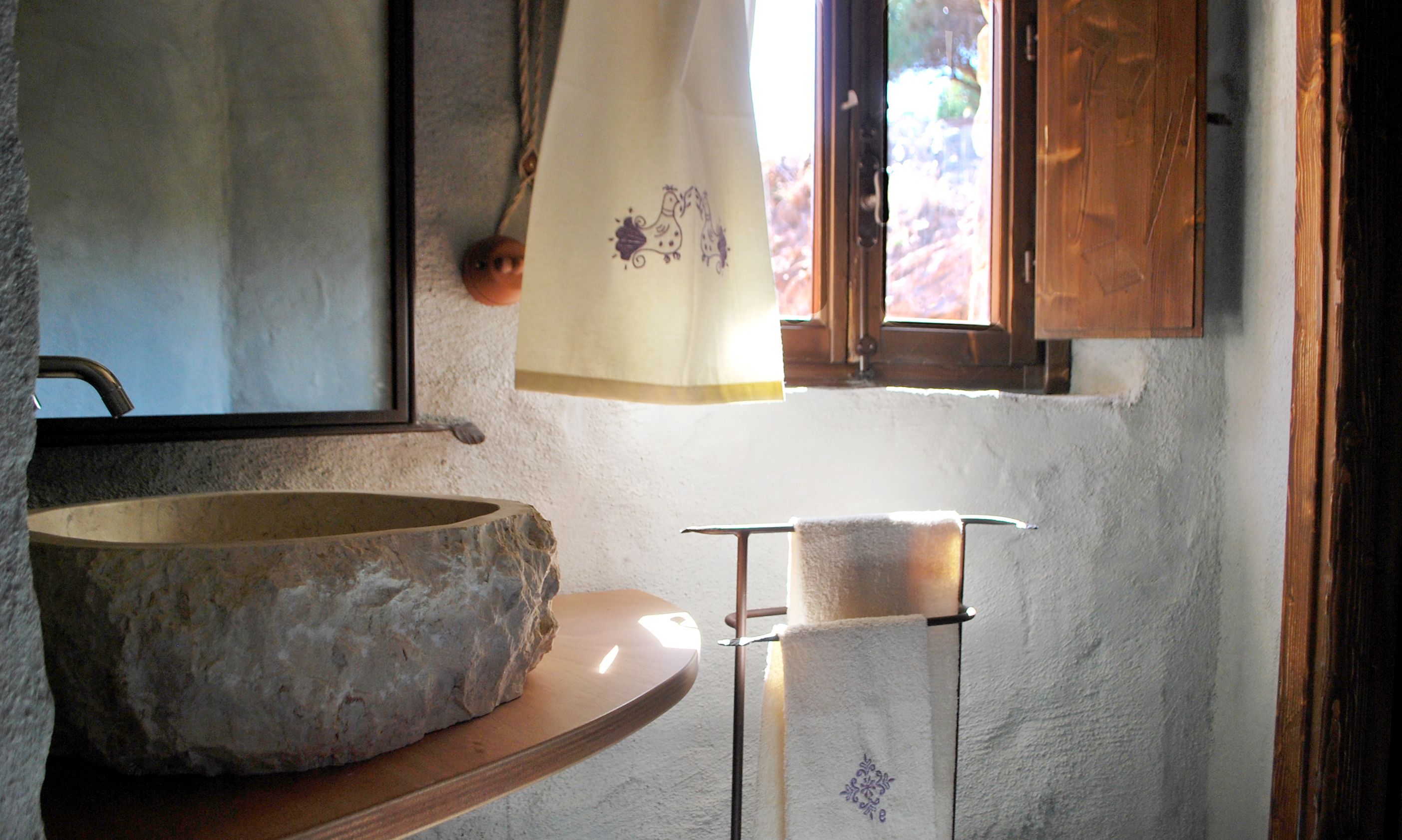 A rustic bathroom with a stone basin, a wooden shelf, and a window with a curtain.