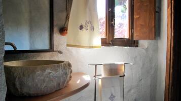 A rustic bathroom with a stone basin, a wooden shelf, and a window with a curtain.