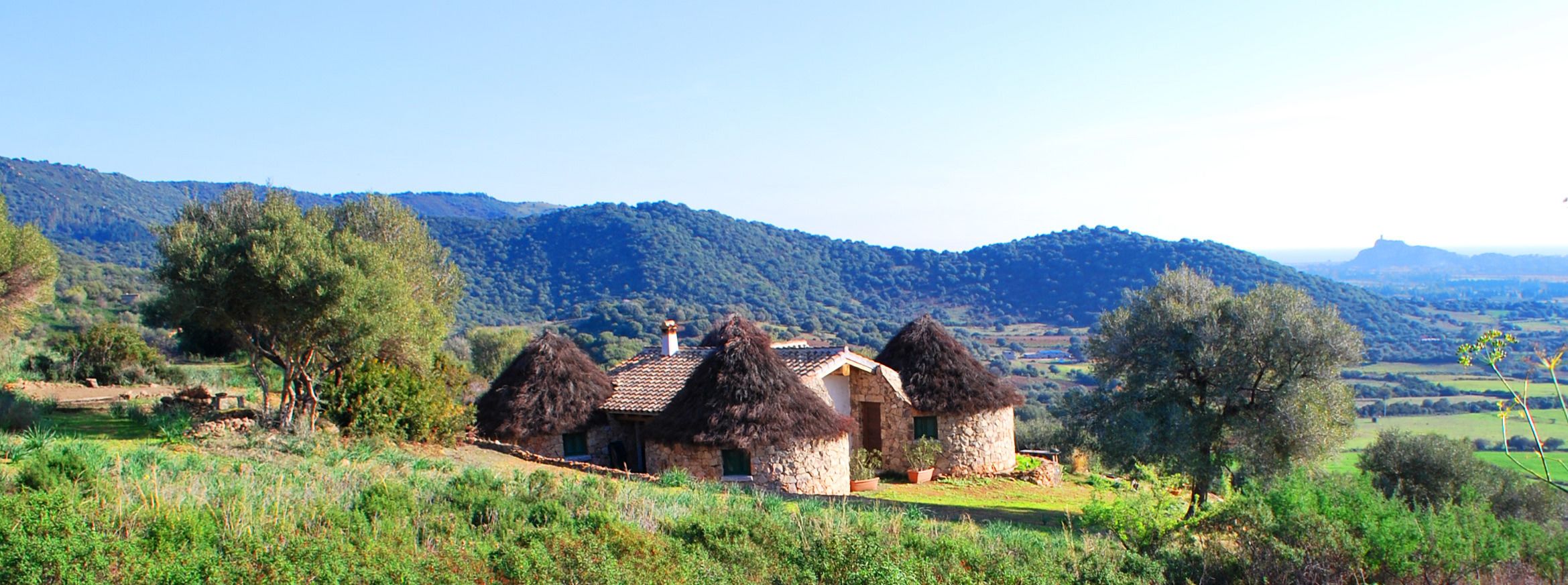 A rustic stone house with thatched roofs, surrounded by lush greenery and hills in the background.
