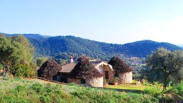 A rustic stone house with thatched roofs, surrounded by lush greenery and hills in the background.